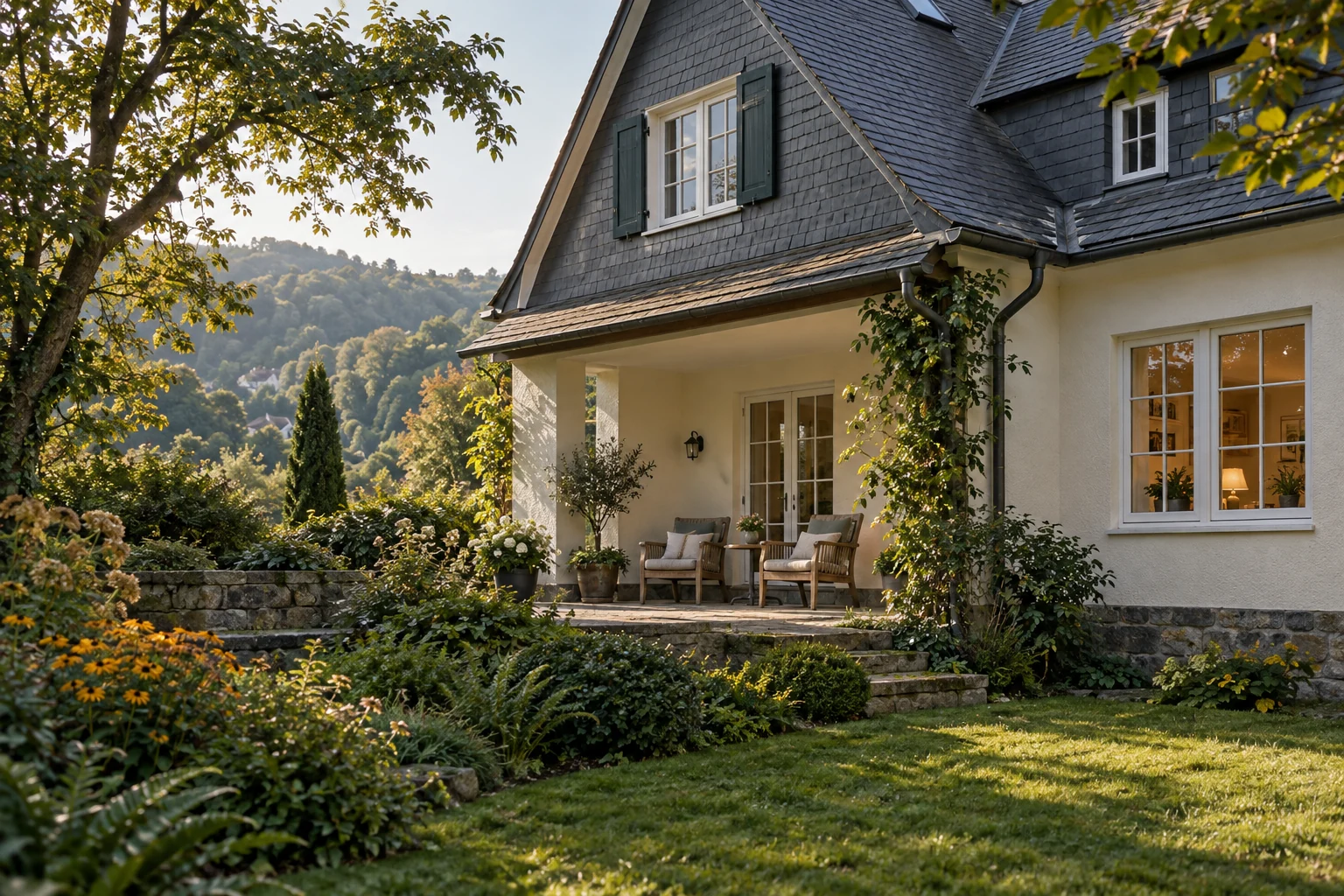 Ein Haus mit Schiefergiebel und geschützter Terrasse mit zwei Sesseln in einem grünen, hügeligen Wohnumfeld.